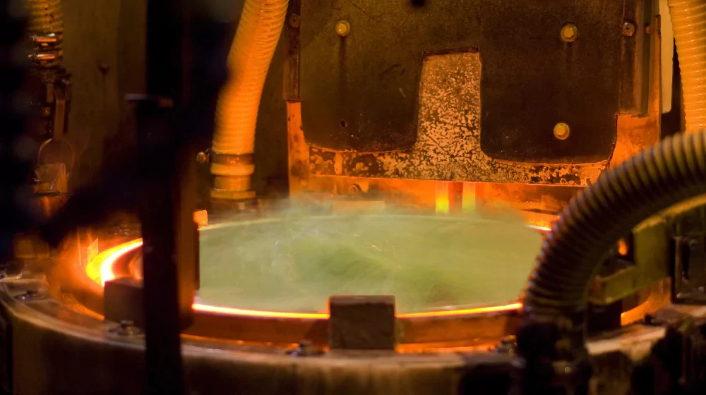 Glowing molten metal in an industrial furnace, surrounded by dark machinery and corrugated hoses