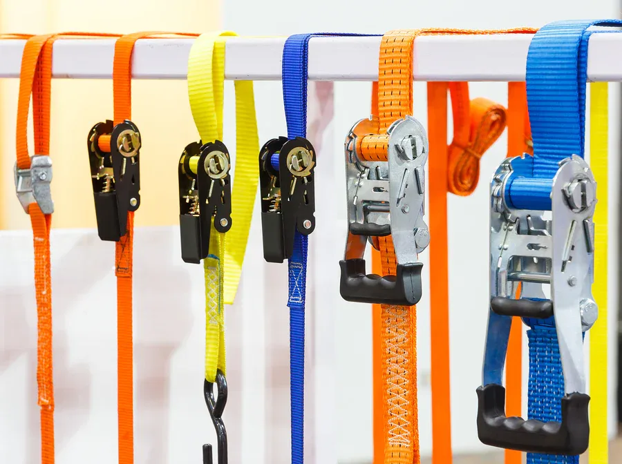 Orange, yellow, and blue ratchet tie-down lashing straps with metal buckles hanging on a white rail