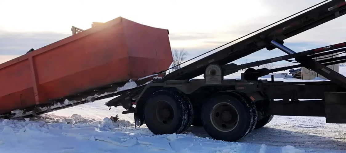 A heavy equipment trailer lifting a red industrial container in a snowy outdoor setting