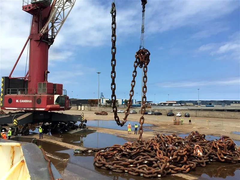 Heavy lifting crane suspended by chains hoisting a pile of rusty anchor chain links in an industrial yard