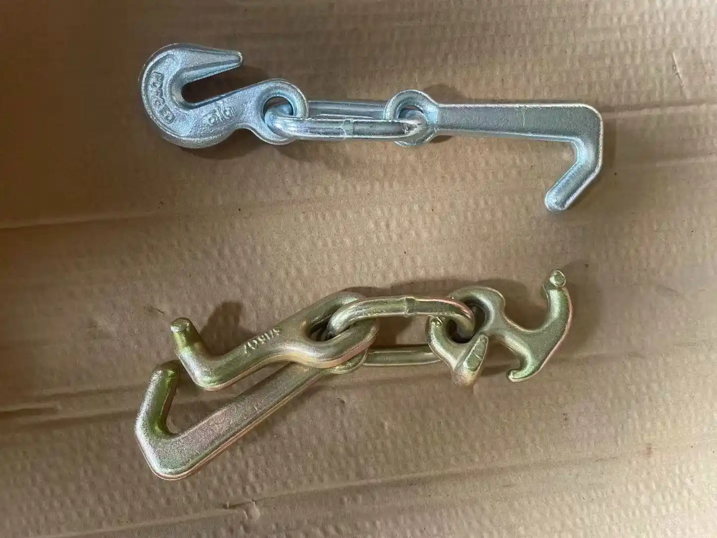 Two forged steel binder hook assemblies, one silver and one yellow-zinc, lying on corrugated cardboard