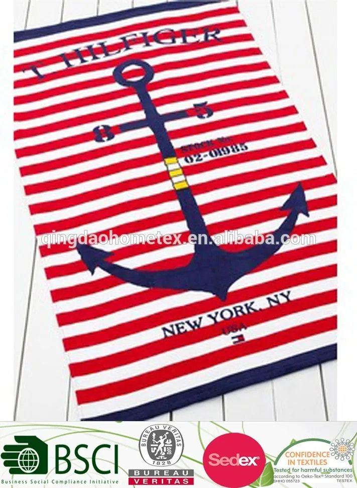 Red and white striped beach towel with navy anchor print and brand lettering, lying on a white wooden surface