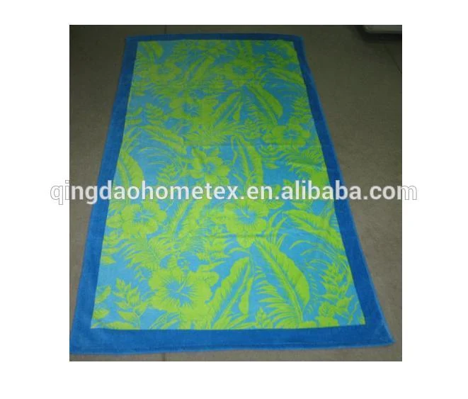 Rectangular beach towel with blue border and green and blue tropical leaf print, laid on a concrete floor