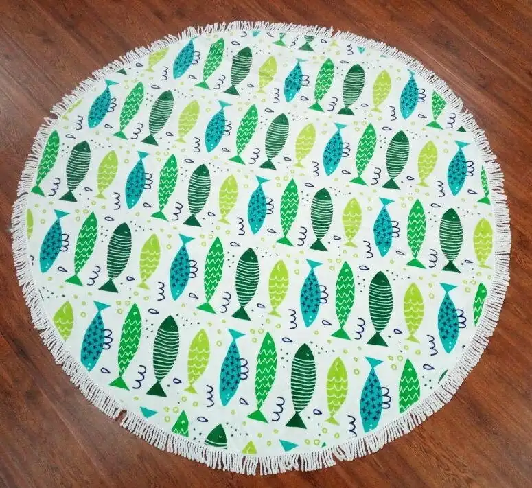 Round white beach towel with fringe and a colorful green and blue fish pattern, laid on a wooden floor