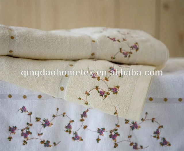 Folded cream cotton towels with purple floral embroidery and dotted ribbon trim stacked on a white towel