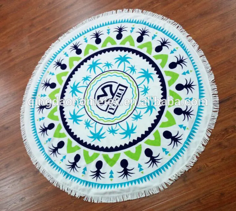 Round white fringed beach towel with blue and green tropical print laid on a wooden floor