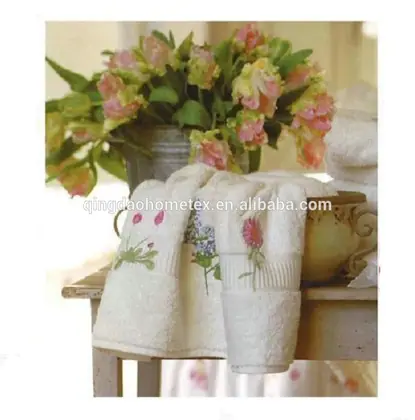 White towels with floral embroidery draped over a table next to a bouquet of pink flowers in a bucket