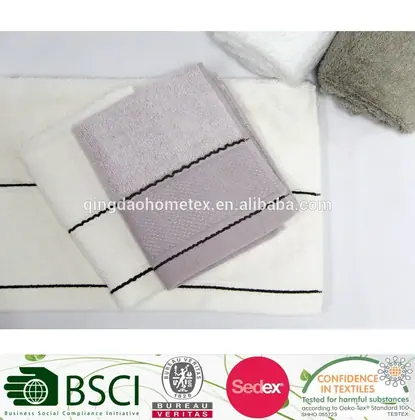 Folded lavender and white terry towels with dark trim stripes, rolled towels at top, certification logos below.