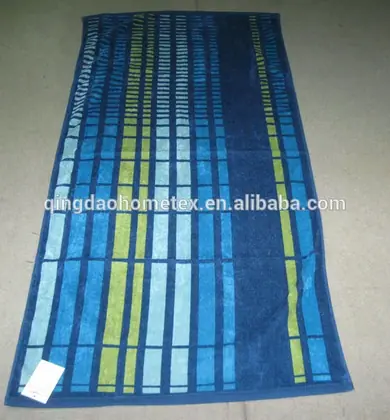 Rectangular woven beach towel with blue, light blue, and green striped geometric pattern laid flat on a gray floor