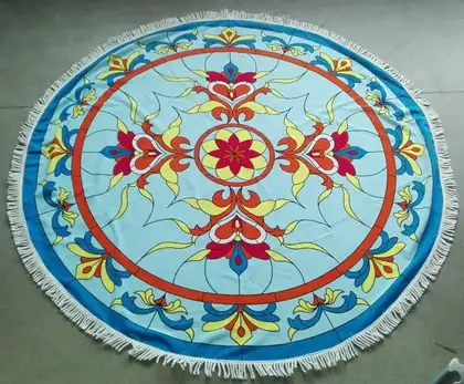 Round blue beach towel with colorful floral mandala print and white fringe, laid flat on a concrete floor