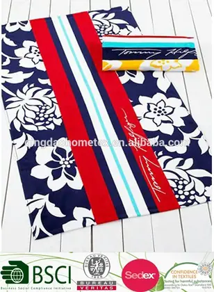 Folded and flat-view beach towels with navy floral print and red, white and blue stripes, with certification logos overlaid at the bottom