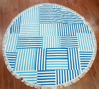 Round beach towel with patchwork of blue and white stripes and fringe, laid on a wooden floor