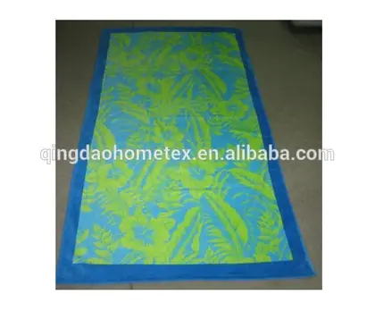 Rectangular beach towel with blue border and green and blue tropical leaf print, laid on a concrete floor
