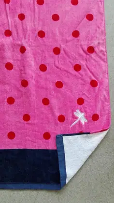 Pink beach towel with red polka dots and navy border, folded corner revealing white reverse side on grey ground