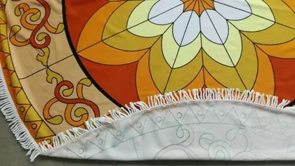 Close-up of a round orange and yellow patterned fabric item with white fringe, partially folded on a grey surface