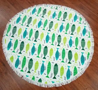 Round white beach towel with fringe and a colorful green and blue fish pattern, laid on a wooden floor