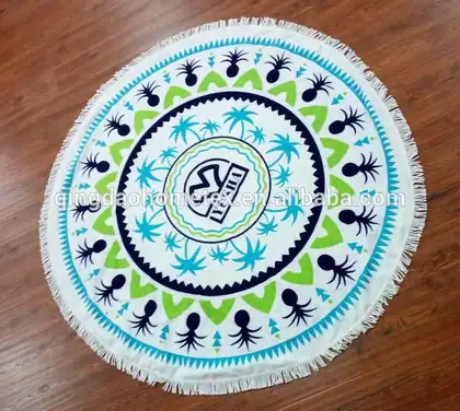 Round white fringed beach towel with blue and green tropical print laid on a wooden floor