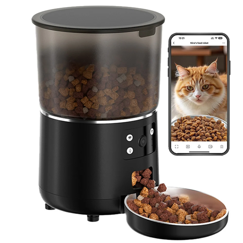 Black automatic pet feeder dispensing kibble into a metal bowl next to a smartphone showing a cat