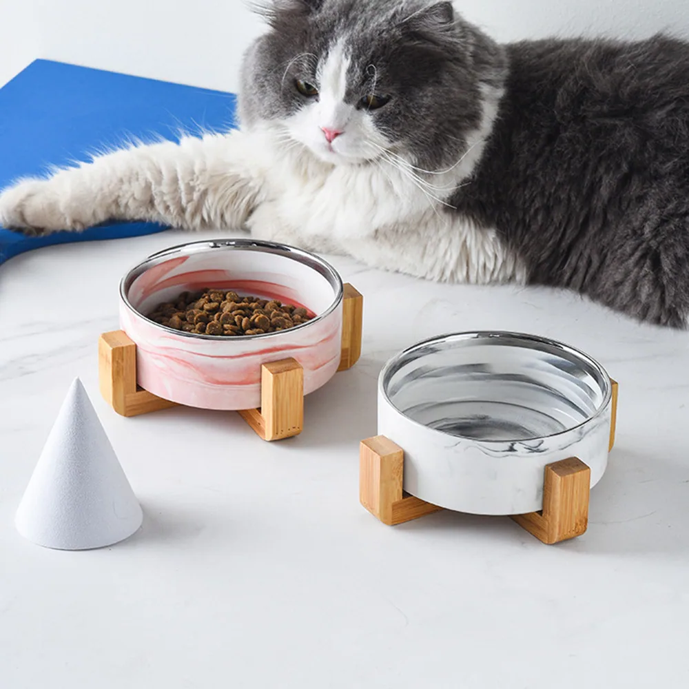 Two elevated marble and wood pet bowls, one pink with food and one white with water, with a cat lying behind them.
