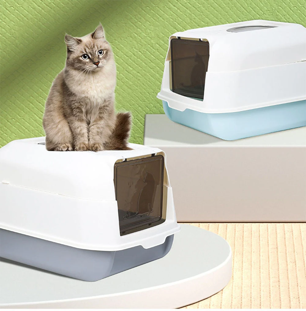 A fluffy cat sits atop a white covered litter box with a grey base, while another white and blue covered litter box sits on a stand in the background.