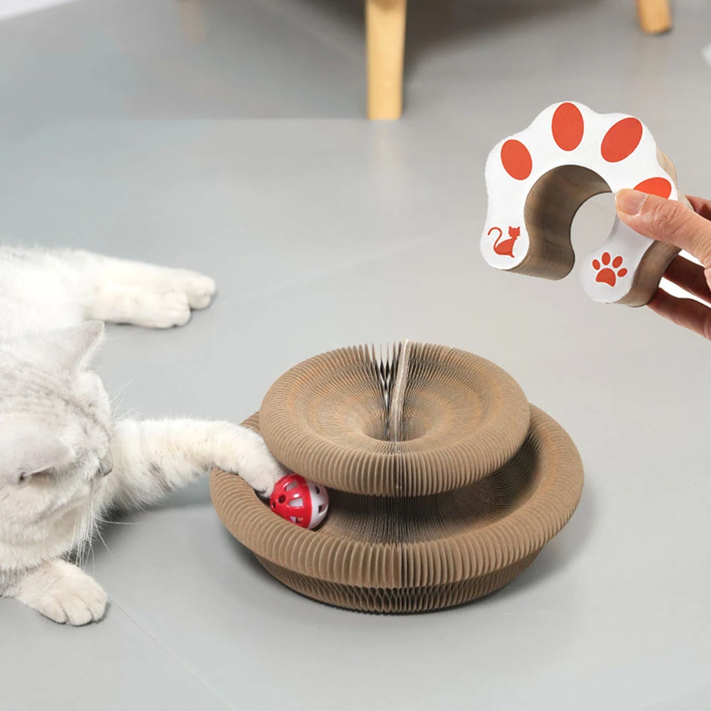 A white cat plays with a layered cardboard scratcher toy containing a red ball, while a hand holds a paw-shaped accessory.