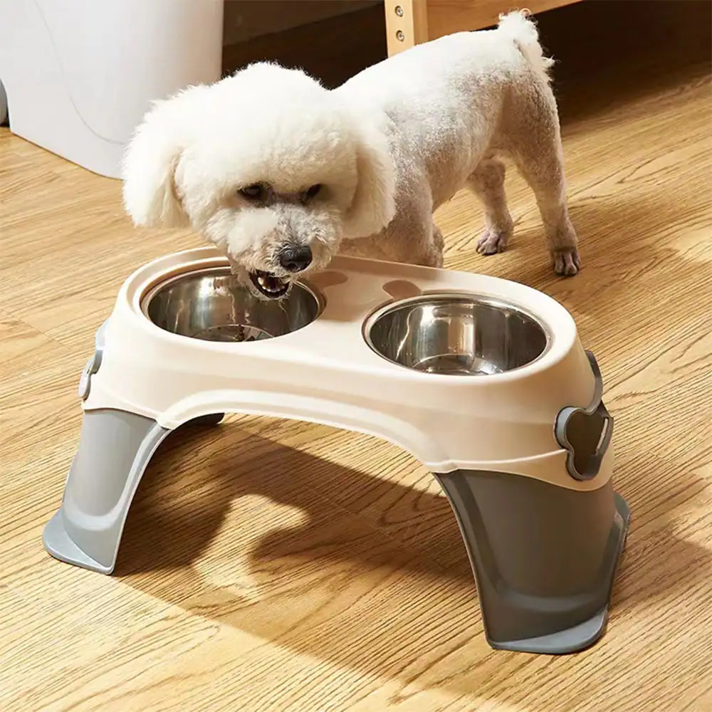 A white dog eating from a raised beige and grey plastic feeder with two stainless steel bowls.