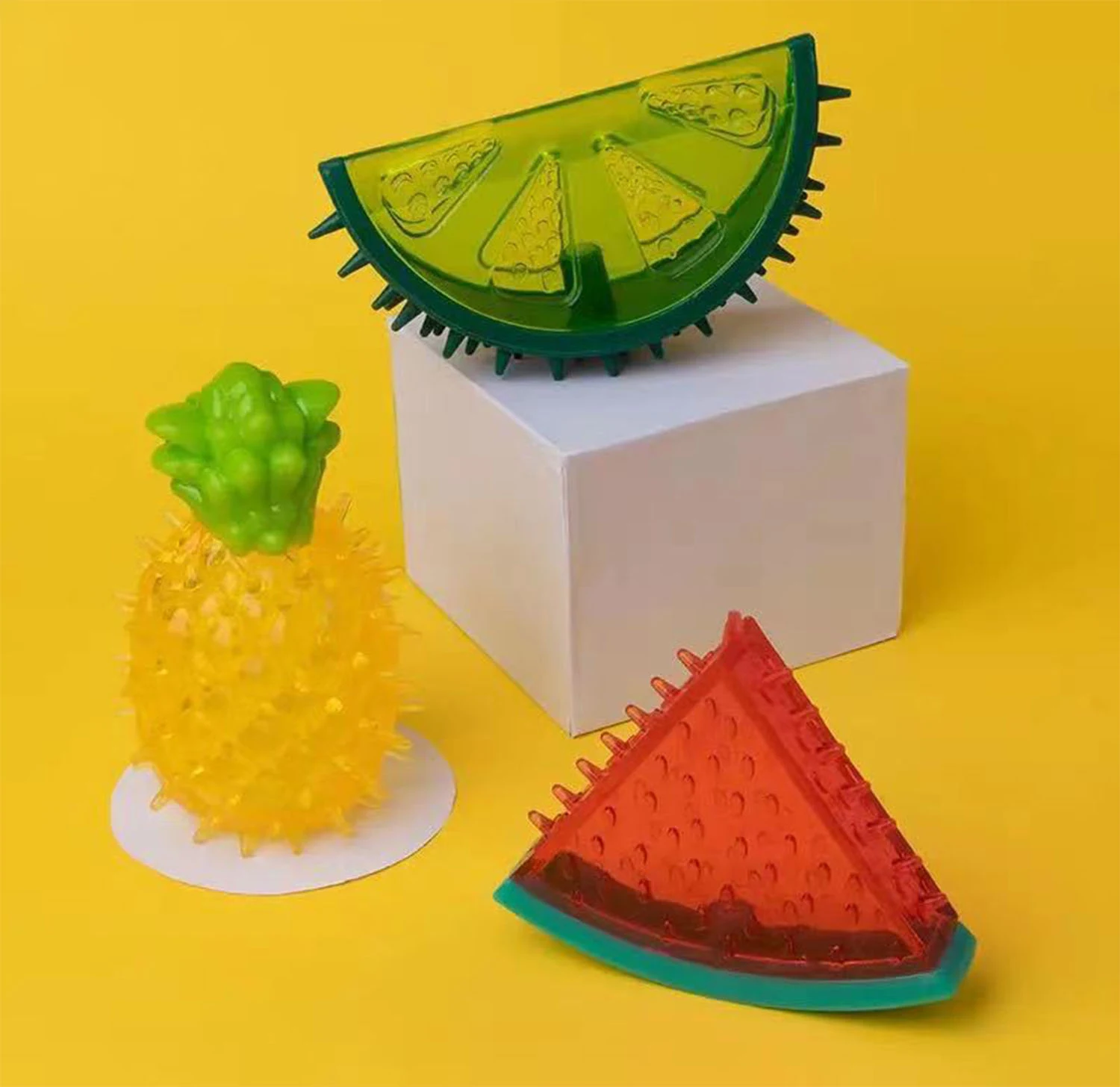 Translucent green and red fruit slice pet toys and a yellow pineapple toy with green top arranged on yellow background