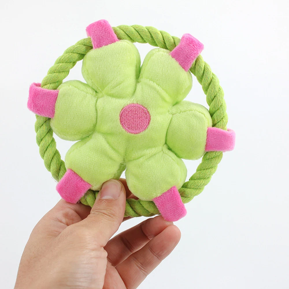 A hand holds a green and pink plush flower dog toy attached to a rope ring against a white background.