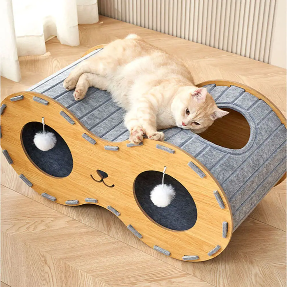 A light-colored cat resting on a wooden and felt cat house with hanging pom-pom toys.