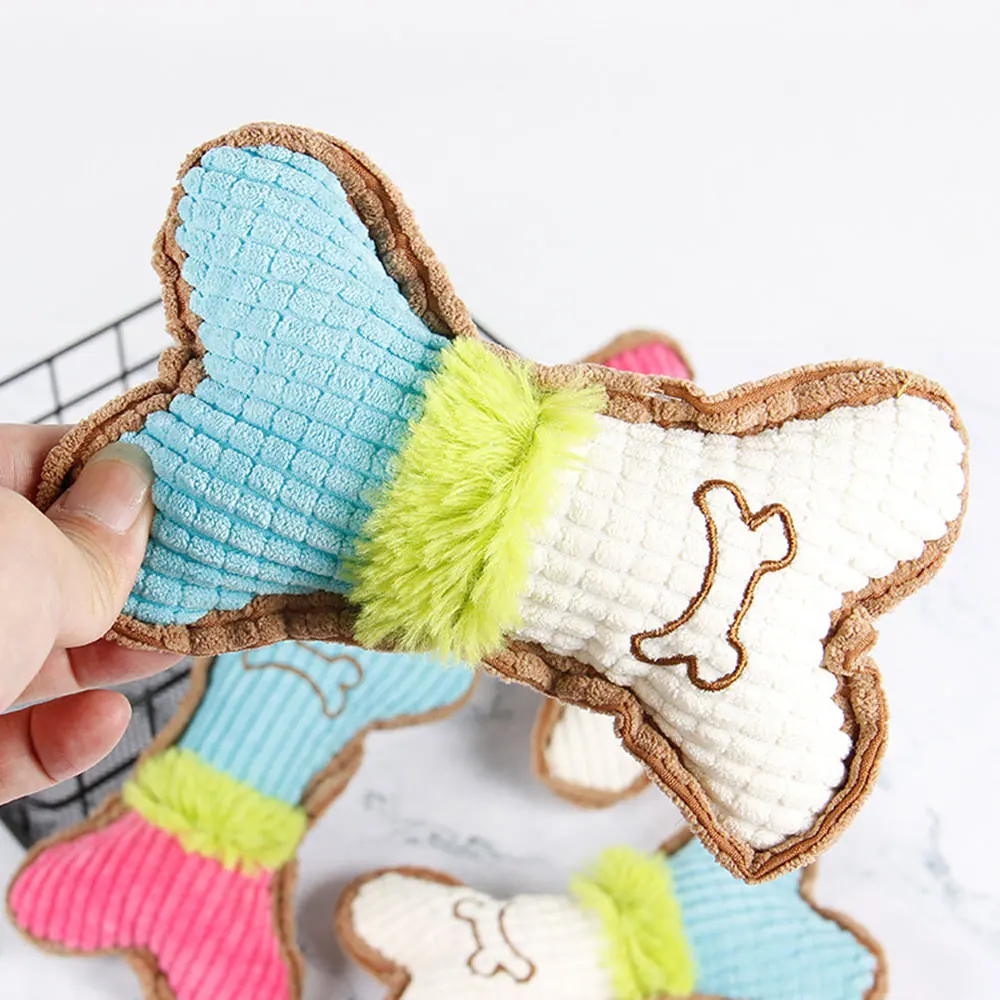A hand holds a bone-shaped dog chew toy featuring blue, white, and green fabric with a pink tail.