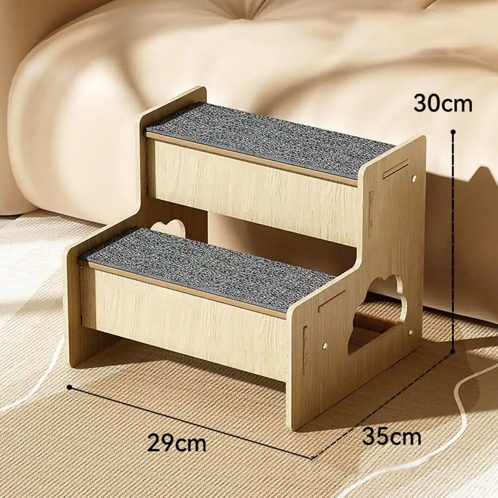 Two-step wooden pet stairs with grey carpeted treads and side handles, placed on a rug in front of a beige sofa.