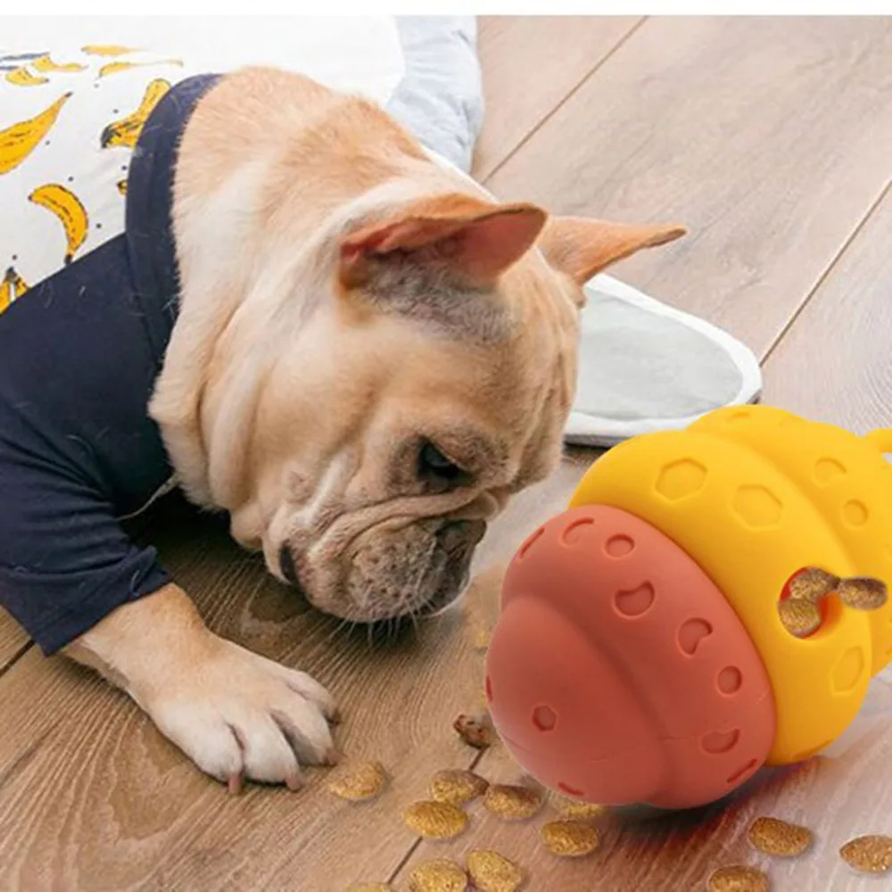 A French bulldog wearing a shirt looks at a yellow and red puzzle toy dispensing treats on a wooden floor.