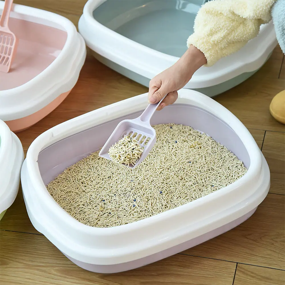 A hand uses a scoop to clean a large, open white and purple cat litter box filled with pellet litter.