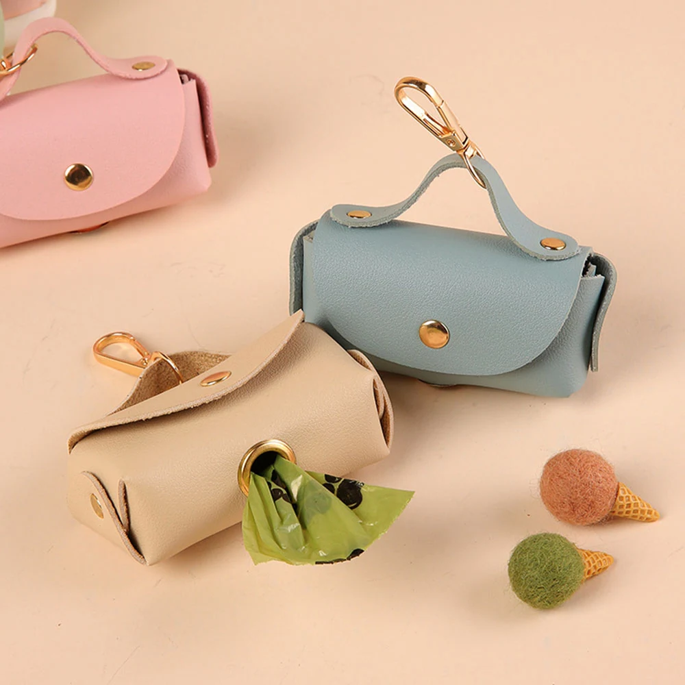 Pink, blue, and beige leather pet waste bag holders with gold hardware, one dispensing a green bag.