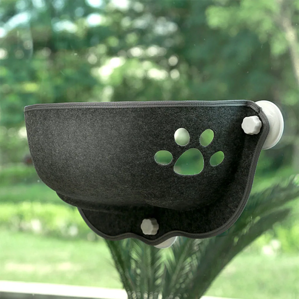 A black fabric cat bed with paw print cutouts attached to a window with suction cups.
