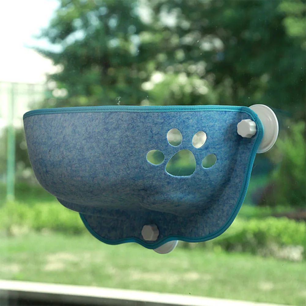 A blue felt pet hammock with paw print cutouts is mounted to a window using white suction cups.