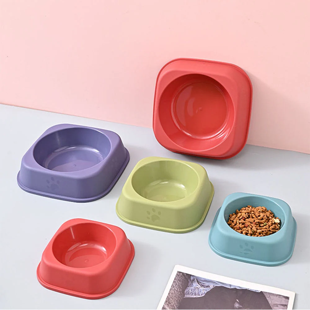 Five colorful square plastic pet bowls arranged on a white surface with a pink background.