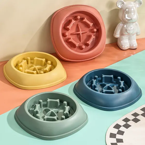 Four colorful slow feeder dog bowls in yellow, red, blue, and green arranged on a multi-colored surface with a decorative bear statue.