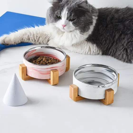 Two elevated marble and wood pet bowls, one pink with food and one white with water, with a cat lying behind them.