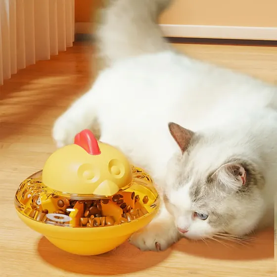 A white cat lying on a wooden floor next to a yellow chick-shaped interactive food dispenser toy.