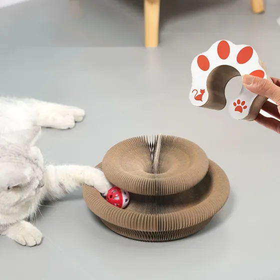 A white cat plays with a layered cardboard scratcher toy containing a red ball, while a hand holds a paw-shaped accessory.