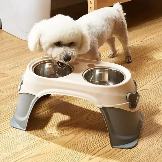 A white dog eating from a raised beige and grey plastic feeder with two stainless steel bowls.