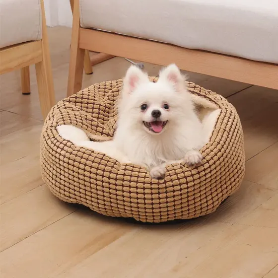 A small white dog resting comfortably inside a round, beige, corduroy-textured pet bed.