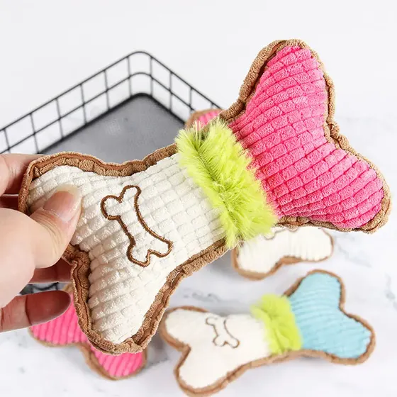Hand holding a bone-shaped pink and white plush dog toy with a green fuzzy center, with similar toys in the background.