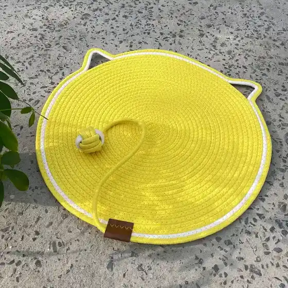 A bright yellow, cat-shaped woven rope mat with a ball toy attached on a concrete floor.