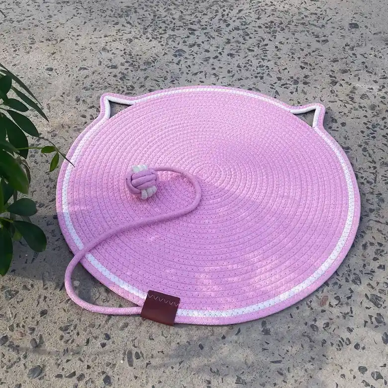 A pink woven cat-shaped mat with a tail and knot, placed on an outdoor concrete floor next to a plant.