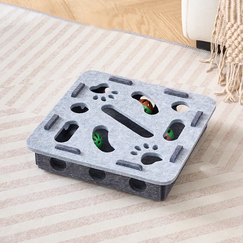 A grey square puzzle box with felt top and cutouts revealing balls inside, sitting on a rug.