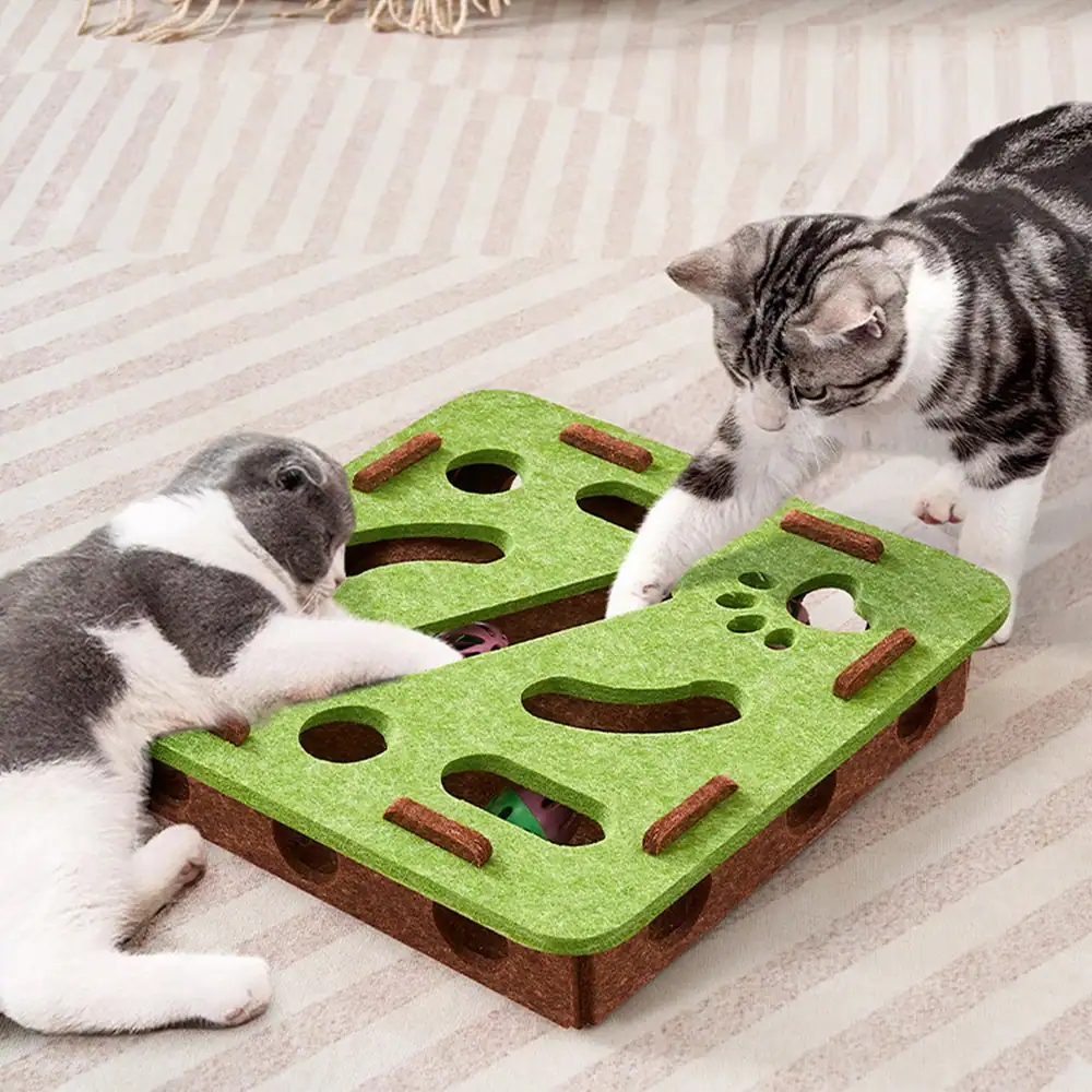 Two cats playing with a green and brown felt puzzle toy on a striped rug.