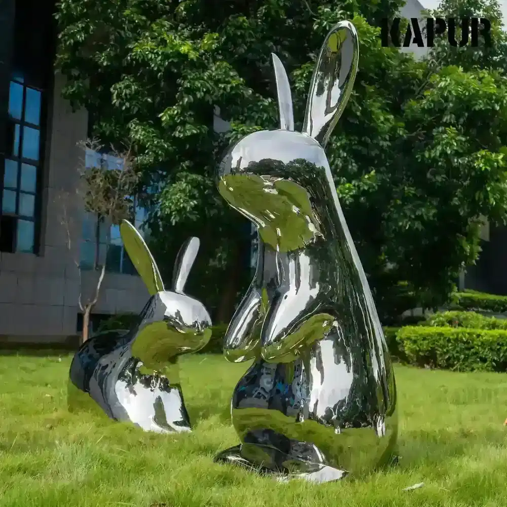 Polished stainless steel rabbit sculpture standing on a paved outdoor plaza, reflecting the sky and surrounding greenery
