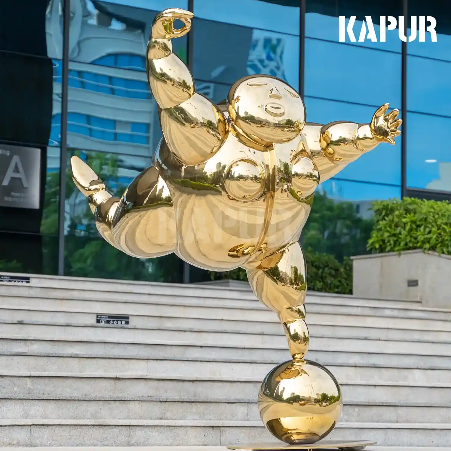 Abstract stainless steel sculpture of a dancing fat man standing on a paved outdoor plaza, reflective surface catching sunlight